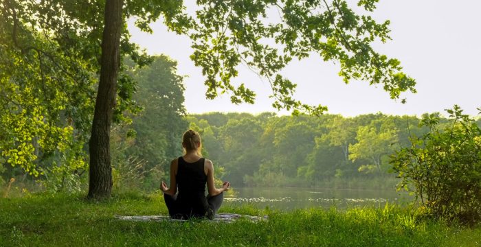 Back view of unrecognizable woman practicing yoga in summer near river outdoors. Rear view of female sitting in meditation pose on yoga mat in lotus position, raising hands up, doing stretching exercise outside